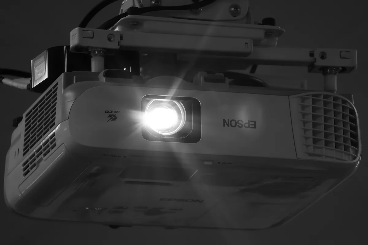 Proyector Epson blanco colgado del techo con un soporte metálico, con la lente encendida