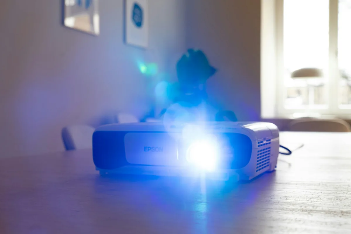 Proyector Epson encendido sobre una mesa de madera en un salón luminoso, con la luz de la lente en azul